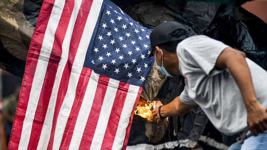 /images/noticias/Um manifestante incendeia uma bandeira dos Estados Unidos pendurada em uma estatua de papel.jpg